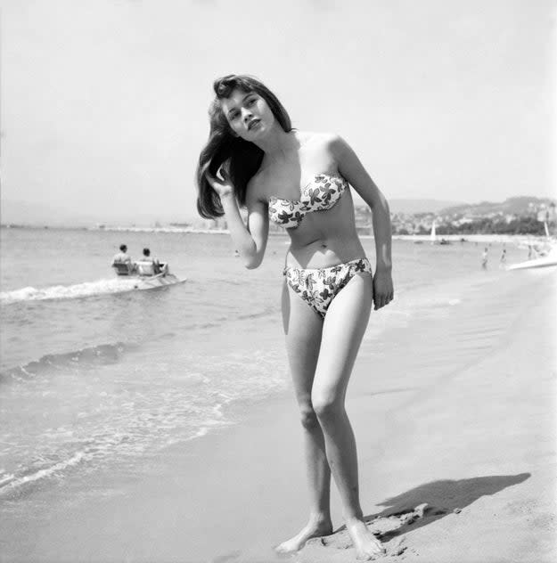 Brigitte Bardot in a floral bikini stands on a sandy beach, with a hand in her hair. People are in the background near the water