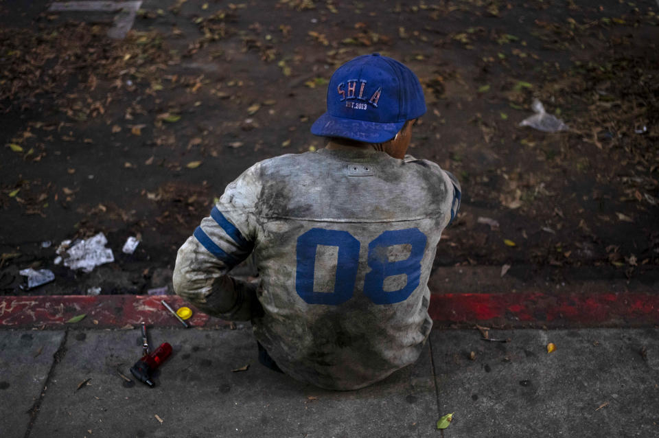 A homeless man wearing a ragged sports jersey passes time on the curb of a street in Los Angeles, Friday, Oct. 20, 2023. (AP Photo/Jae C. Hong)