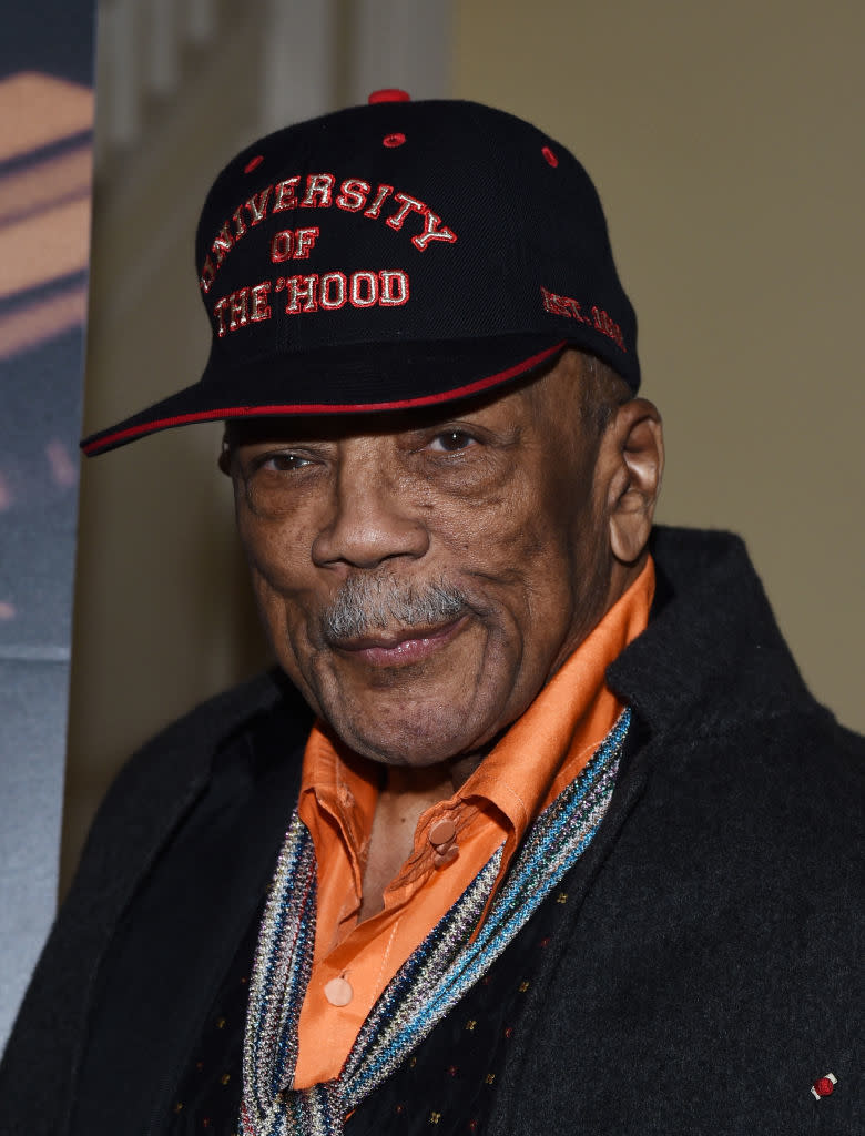 Quincy in a "University of the Hood" cap, orange shirt, and tweed jacket