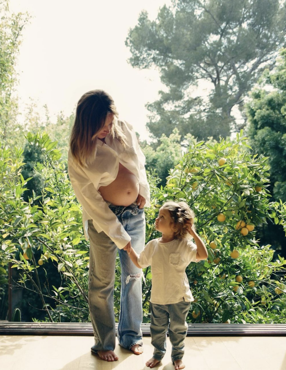 Ashley  lifting shirt to show belly to Jupiter, both smiling, standing indoors by a window with trees outside