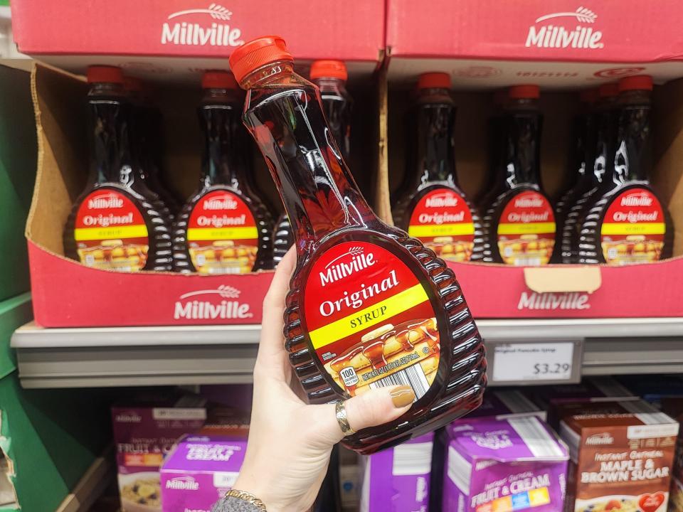 A hand holding a bottle of Millville original syrup in front of a larger display.