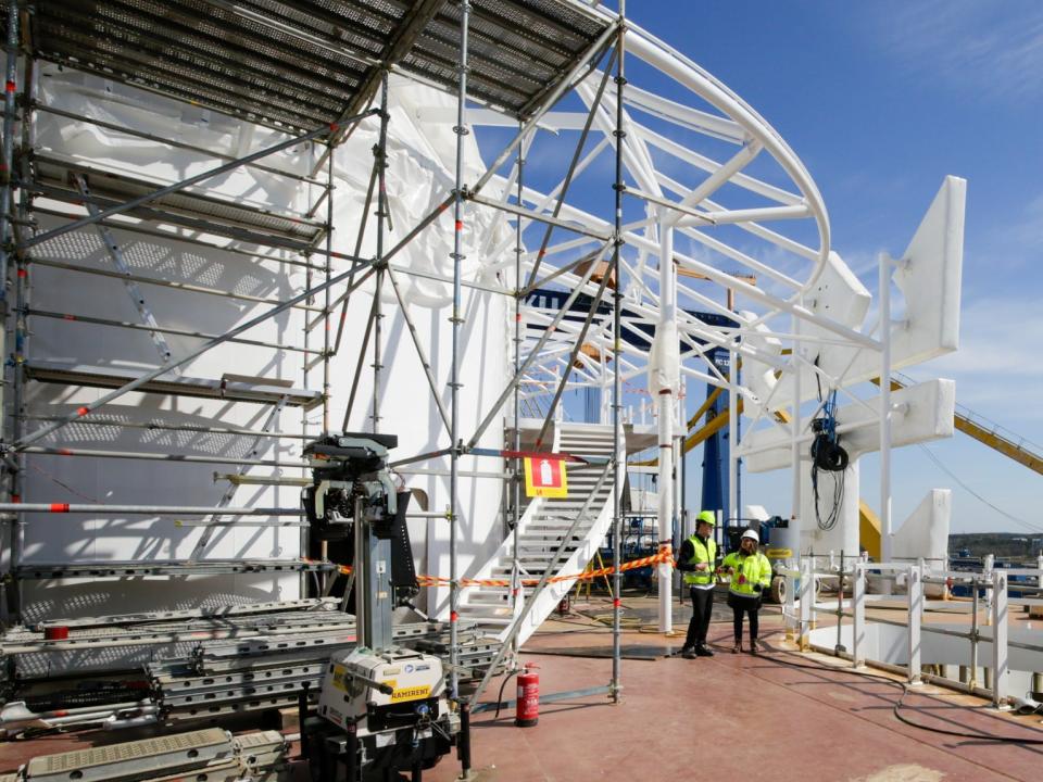 Two people in high-visibility jackets and white hard hats standing on the deck of the Icon of the Seas that is under construction.