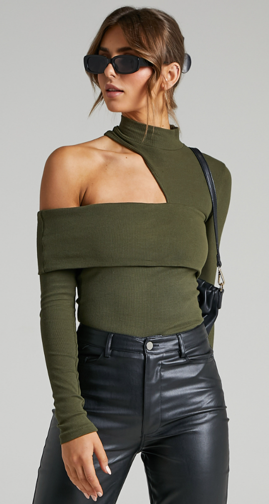 A woman stands in olive green top with shoulder cut out, black sunglasses, bag and leather pants