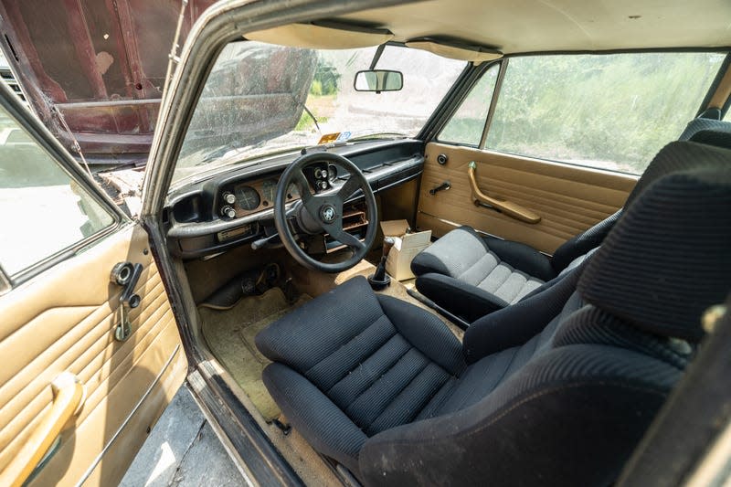 a photo of the tan interior of a long-abandoned 1974 bmw2002tii. the carpet and door upholstery is faded tan; the seats, steering wheel and dashboard are all faded black. there is random trash in the car, which has sat abandoned in a florida field for 25 years