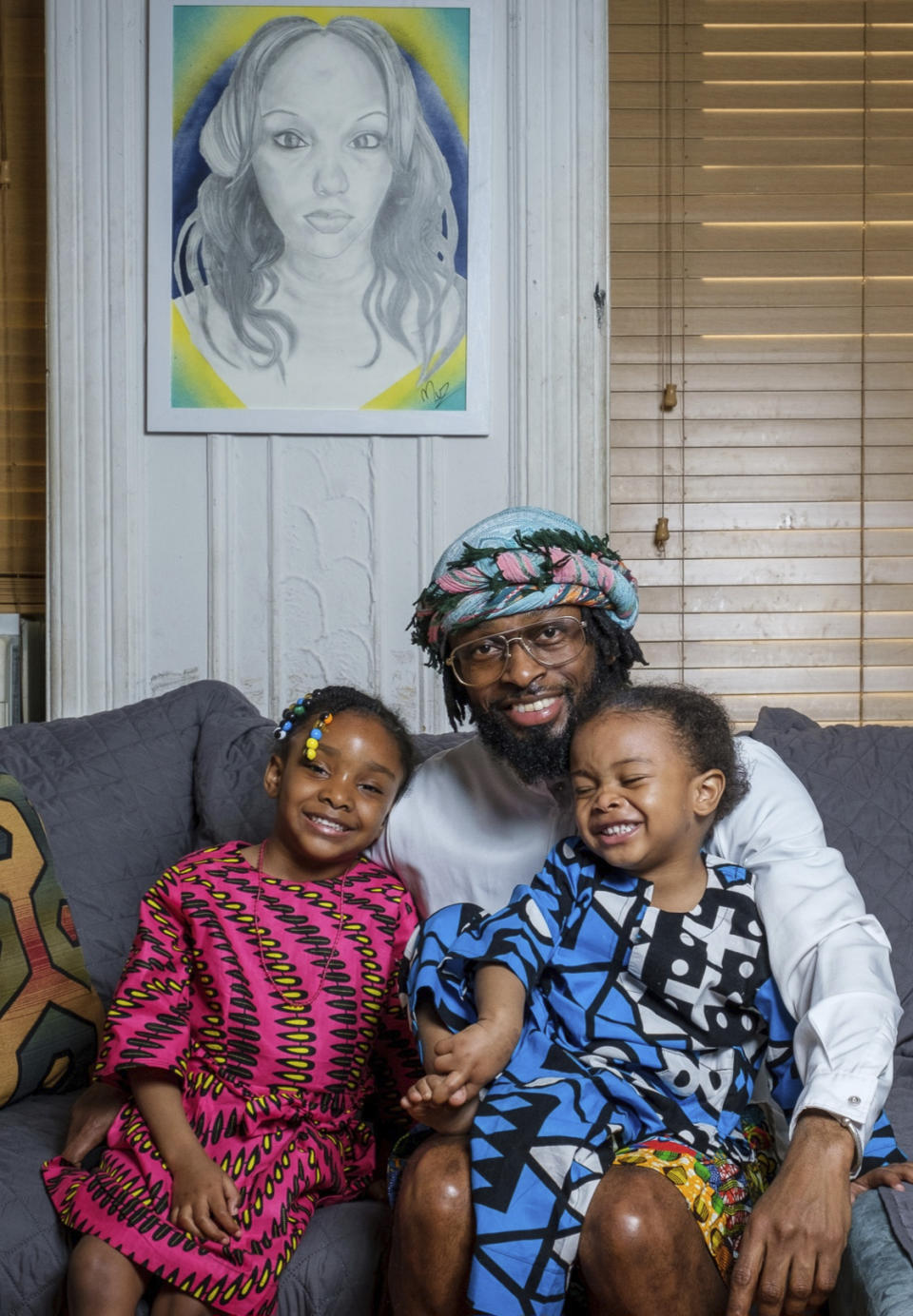 In this photo provided by Henry Danner, Omari Maynard sits with his children, Anari, left, and Khari, next to a painting of their late mother, Shamony Gibson, at home in the Brooklyn borough of New York on April 9, 2022. Gibson passed away in 2019, two weeks after giving birth to Khari due to a pulmonary embolism. “She wasn’t being heard at all,” said Maynard, an artist who now does speaking engagements as a maternal health advocate. (Henry Danner via AP)