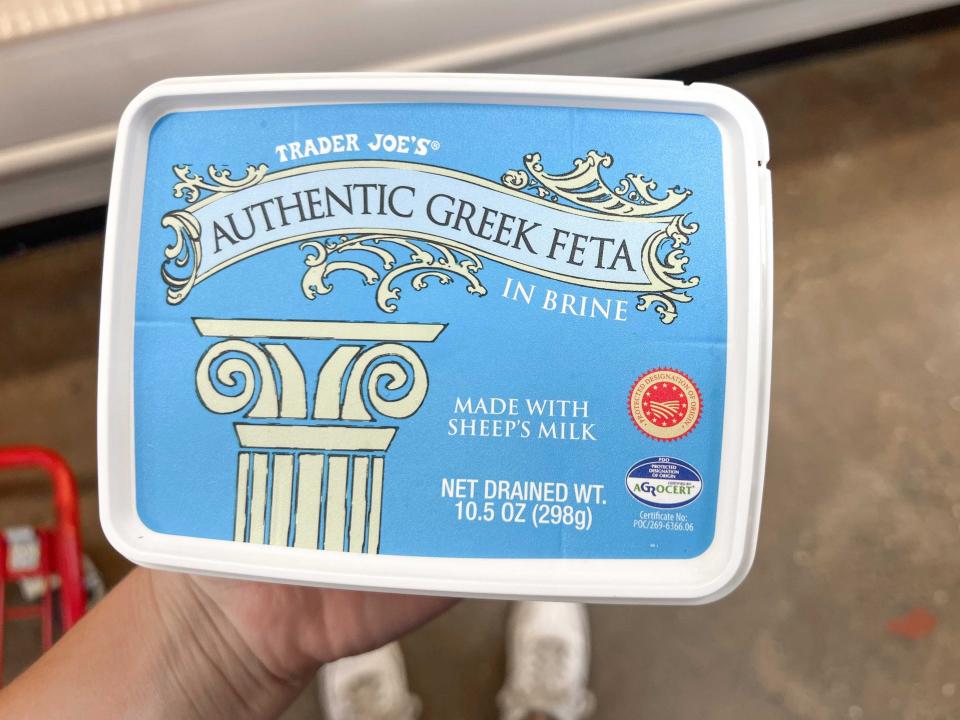 A hand holds a blue container of feta with an illustration of a Greek-style pillar on the lid