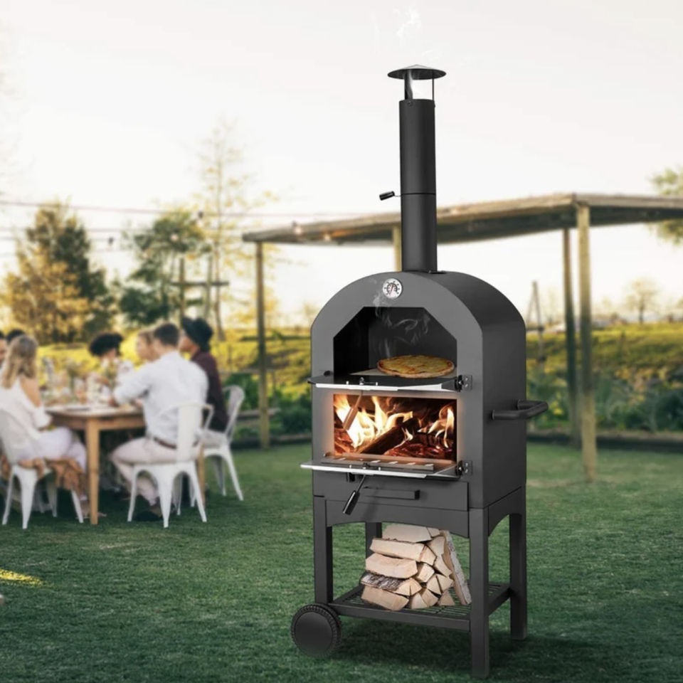 Steel Freestanding Wood-Fired Pizza Oven in black on green grass with people eating in background (Photo via Wayfair)
