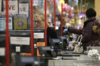A cashier at a grocery store works while standing behind a plastic shield, Thursday, March 26, 2020, in Quincy, Mass. Grocery stores across the U.S. are installing protective plastic shields at checkouts to help keep cashiers and shoppers from infecting each other with the coronavirus. The new coronavirus causes mild or moderate symptoms for most people, but for some, especially older adults and people with existing health problems, it can cause more severe illness or death. (AP Photo/Steven Senne)
