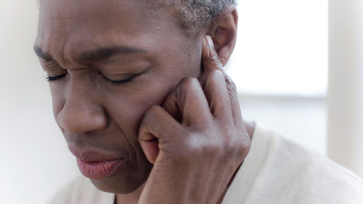  Close up of a woman's face as she winces and holds a finger to one ear, as if trying to tune out a noise. 