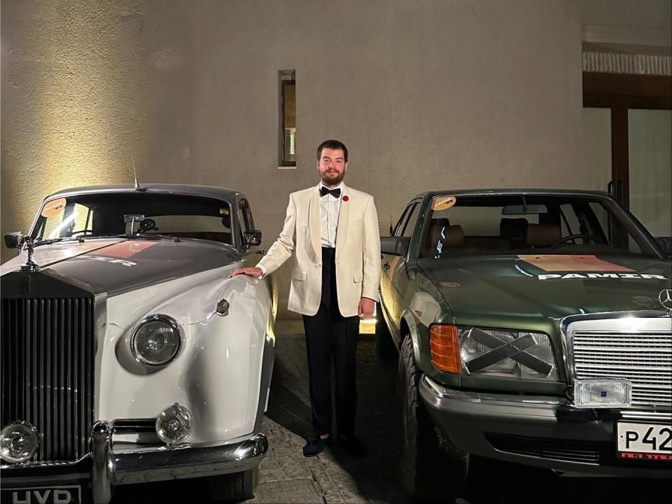 A man in a white dinner jacket standing with two vintage cars.
