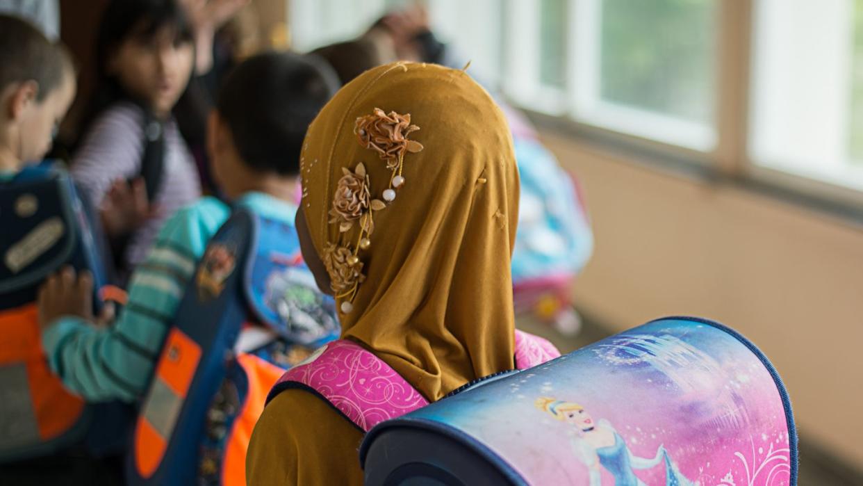 Das Symbolfoto zeigt ein Mädchen mit Kopftuch vor einem Klassenzimmer.