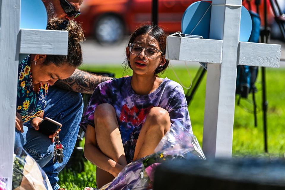 A girl sits in front of a cross holding her knees and crying.