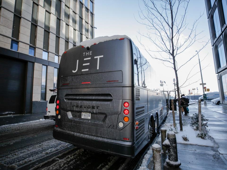 A matte black bus that reads "The Jet" on the side. Passengers with bags are boarding the bus or putting their bags away into the lower storage compartment.A matte black bus that reads "The Jet" on the side. Passengers with bags are boarding the bus or putting their bags away into the lower storage compartment.