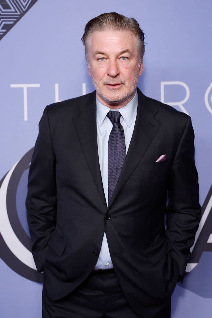 Alec Baldwin in a black suit with tie and pocket square, hands folded, at an event