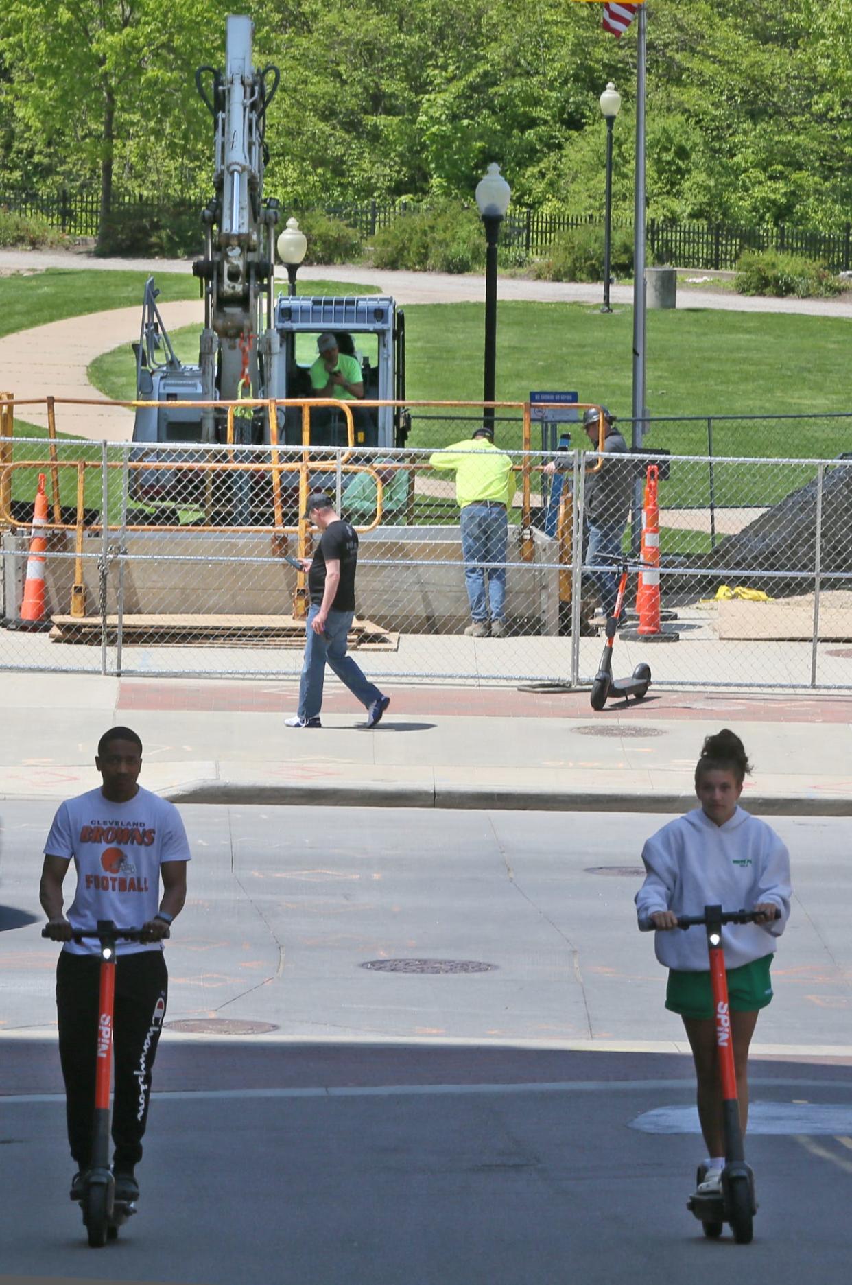 Scooter riders make their way up University Avenue as construction workers install a chilled water line into the O'Neil's building at the entrance to Lock 3 in Akron on Tuesday. Construction on the park will start in a few weeks.