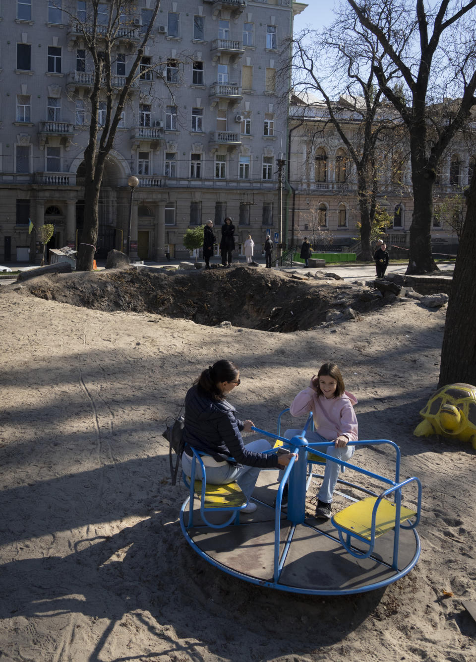 Mother and daughter play in a playground near a huge crater, left by a Russian rocket a week ago, in central Kyiv, Ukraine, Sunday, Oct. 16, 2022. (AP Photo/Efrem Lukatsky)