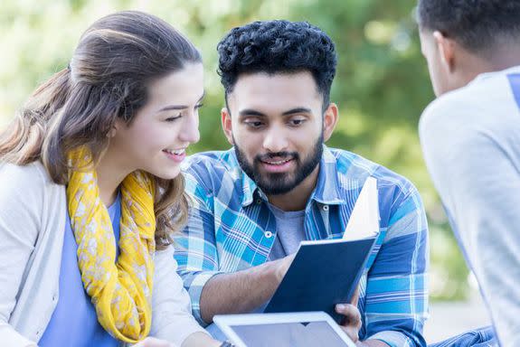 Cheerful Indian male college student shows his female friend something in a book. A male friend is sitting in the foreground. They are studying outdoors on campus on a sunny day.