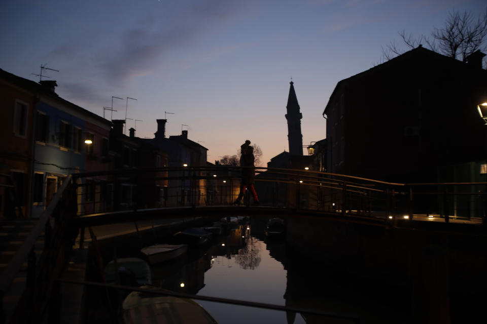In this image taken on Thursday, Jan. 16, 2020, a woman walks over a bridge at sunset, at the Burano island, Italy. The Venetian island of Burano's legacy as a fishing village remains the source of its charms: the small colorful fishermen's cottages, traditional butter cookies that were the fishermen's sustenance at sea and delicate lace still stitched by women in their homes. As the island's population dwindles, echoing that of Venice itself, so too are the numbers of skilled artisans and tradespeople who have kept the traditions and economy alive. (AP Photo/Luca Bruno)