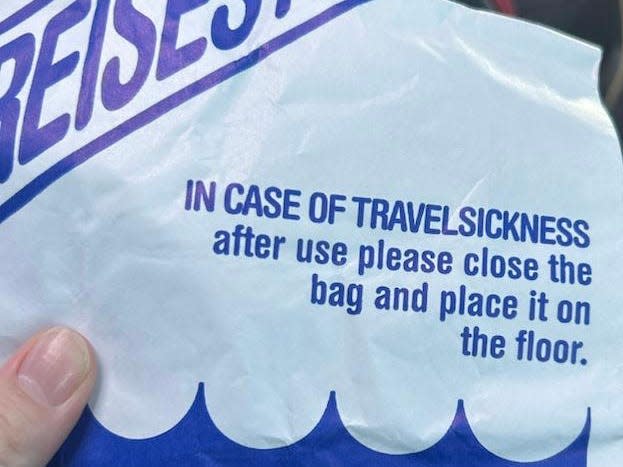 A hand holding a blue and white bag that reads "In case of travelsickness: after use please close the bag and place it on the floor."