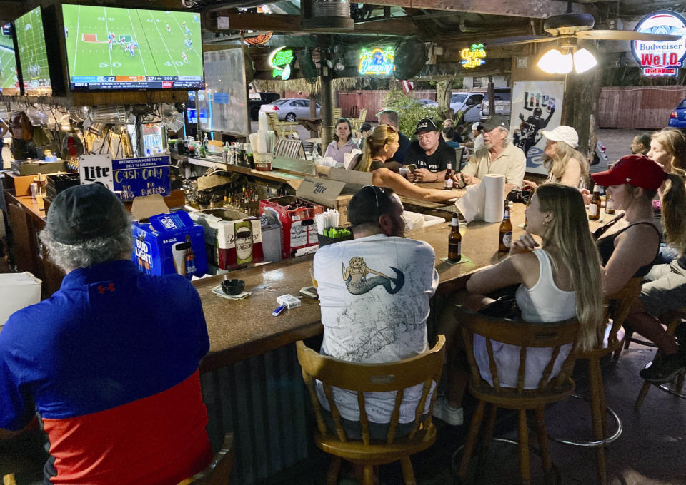 People watch college football games at an outdoor tiki bar under banyan trees in Fort Myers, Fla., on Saturday, Oct. 8, 2022. While some areas are still ravaged by effects of Hurricane Ian, life is returning to normal in other places. (AP Photo/Jay Reeves)