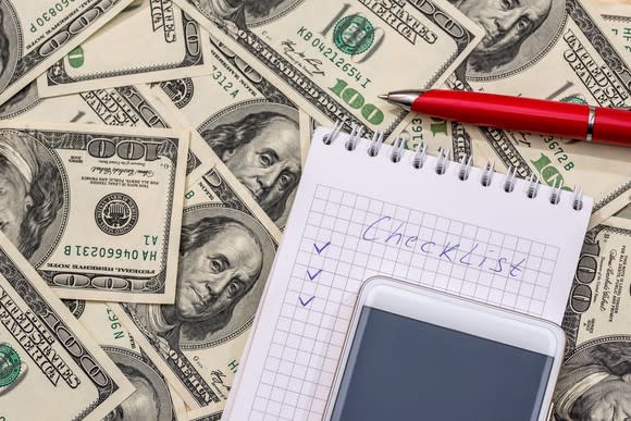 A notebook with a checklist on it with hundred dollar bills in the background.