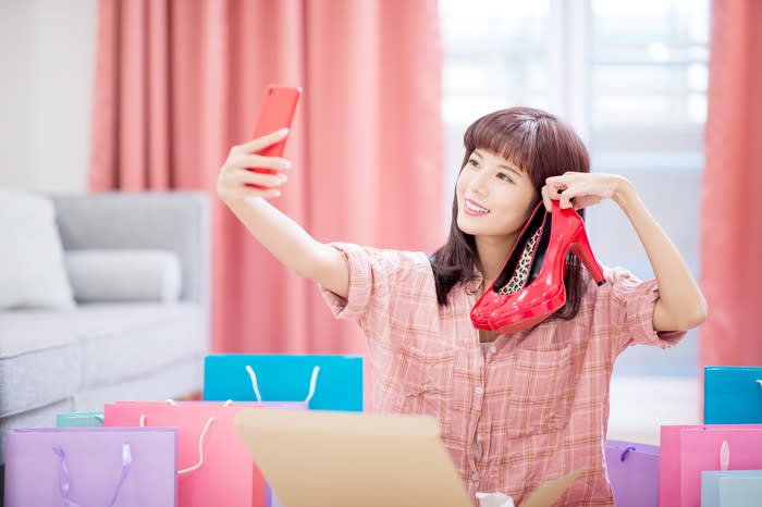 A smiling young woman takes a selfie with a pair of shoes.