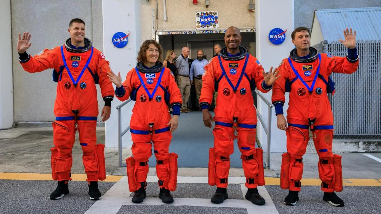  Four astronauts in orange spacesuits waving at the camera in front of a doorway festooned with nasa logos. 