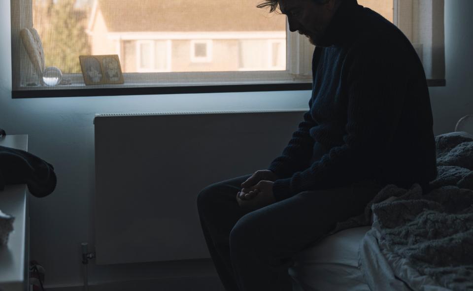 A man is sitting on the edge of a bed in a dimly lit room, looking down with a contemplative expression. The window behind him shows a suburban scene