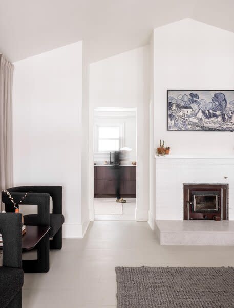 A trove of original details await in the airy living room, including a pitched ceiling, brick fireplace, light-hued hardwood floors, and wraparound windows. An open doorway leads to the kitchen.