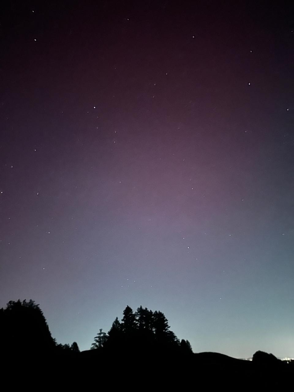 night sky with gradation from white city lights up to blue to pink to black above a dark tree hill silhouette