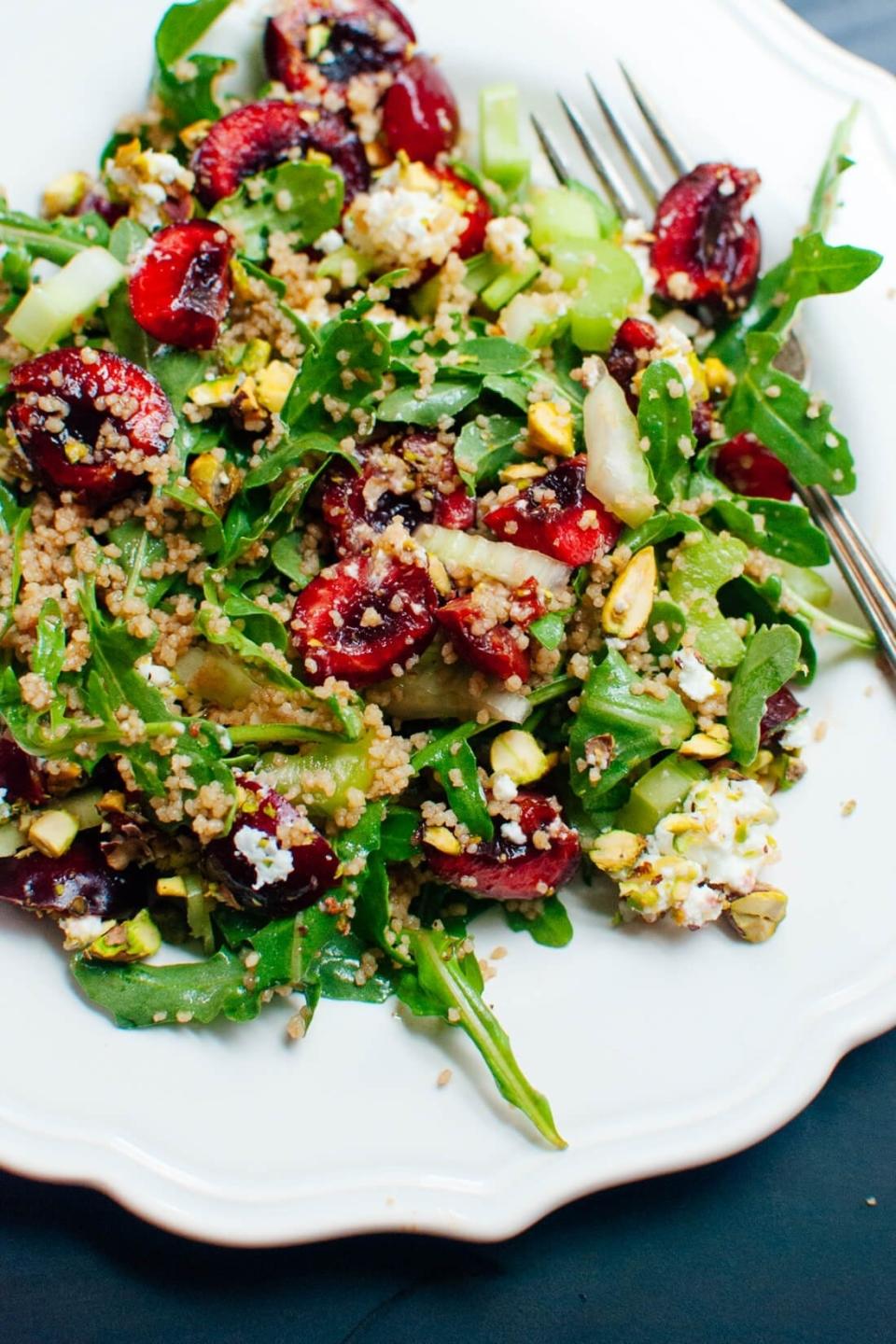 A vibrant salad featuring cherries, quinoa, arugula, goat cheese, and chopped pistachios on a white plate, with a fork placed to the side