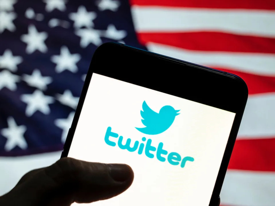 A hand holds a phone with Twitter's logo on the screen in front of an American flag.