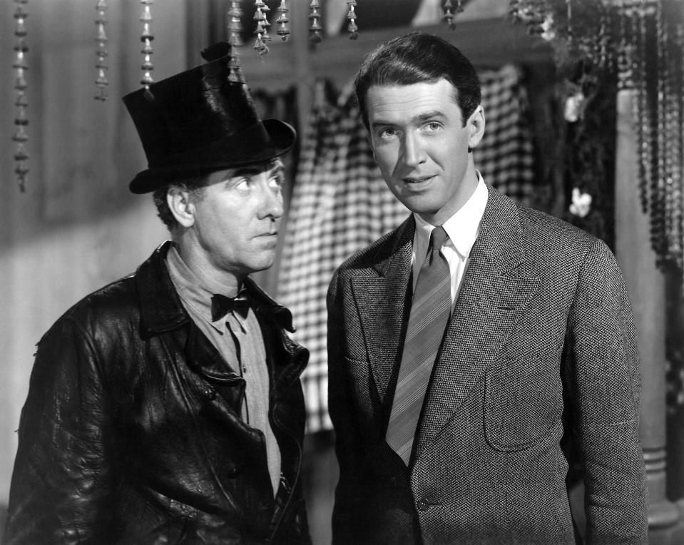 James Stewart in a suit and bowtie stands beside a man with a top hat and leather jacket in a classic film scene