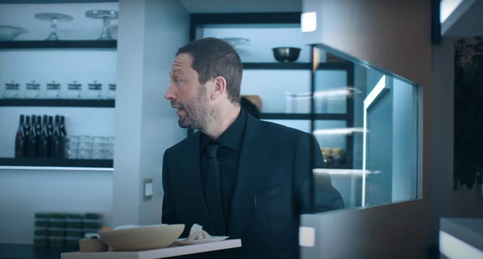 Richie from The Bear wearing a suit enters a modern kitchen, holding a tray with a bowl and a cup. The kitchen features glassware and bottles neatly arranged on shelves