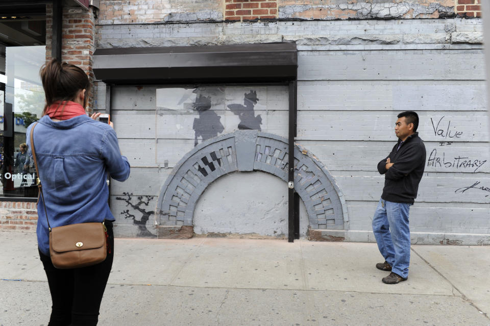 A woman, left, looks at work by British graffiti artist Banksy as Feliciano Perez guards on Saturday, Oct. 19, 2013, in the Brooklyn borough of New York. The building owner has hired security guards and installed a metal gate to protect a work by Banksy. Cara Tabachnick, whose family owns the building, said the goal is to preserve the artwork "so it can be viewed and enjoyed." Most of the Banksy works that have gone up have been tagged over by others, and some have been completely erased. Mayor Michael Bloomberg said last week that graffiti ruins property and is "a sign of decay." (AP Photo/Alyssa Goodman)