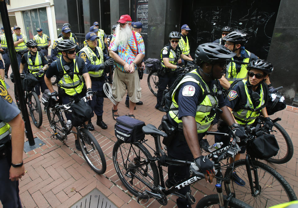 State Police escort local resident, John Miska, red hat, after he was arrested in the locked down downtown area in Charlottesville, Va., Saturday, Aug. 11, 2018. Miska purchased razor blades, which are banned items, in a downtown drugstore. On the the anniversary of white supremacist violence, state and local authorities framed the weekend's heightened security as a necessary precaution. (AP Photo/Steve Helber)