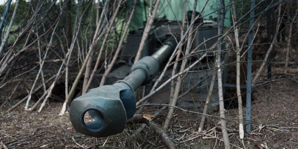 The barrel of a M109A6 Paladin 155mm self-propelled howitzer poking out of fence of sticks while in a camouflaged position in a field.