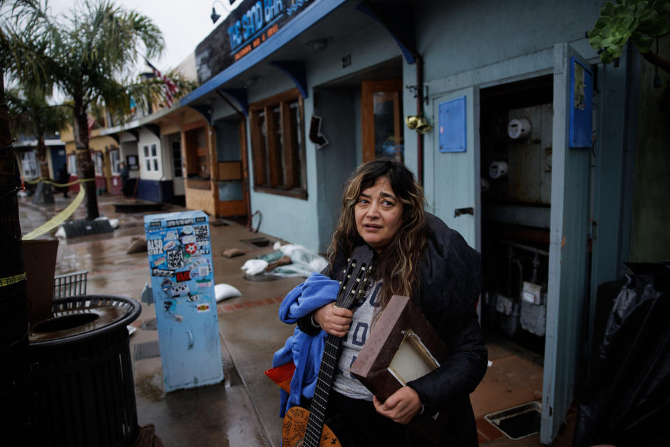 A woman stands outside low bungalows, holding a guitar, a sweatshirt, and a frame.