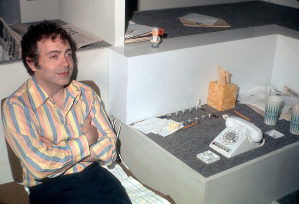 A man sits beside a desk with a rotary phone, notebooks, tissues, and coffee cups. The desk has papers and a small figurine on the elevated section