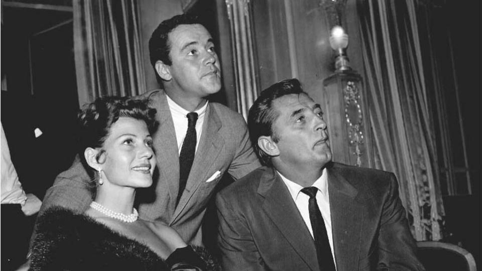 rita hayworth, jack lemmon, and robert mitchum look to the right, hayworth and mitchum are seated next to each other and lemmon stands behind them, she wears a dark fur wrap, triple stranded pearl necklace and pearl earrings, lemmon wears a gray suit and dark tie, mitchum wears a dark suit and tie