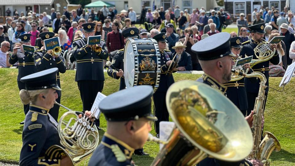 Members of the Band of the RAF Regiment playing shiny brass instruments. The uniforms are grey-blue with gold trim and the band includes a drum with a coat of arms on it.