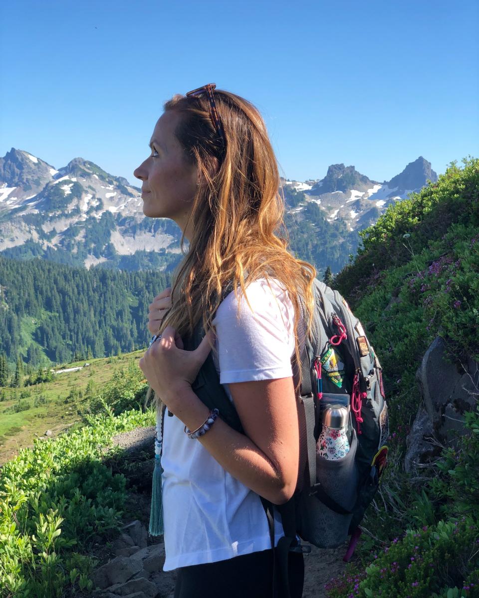 A side-profile shot of Emily wearing a white t-shirt and a hiking backpack in front of mountains and trees. 