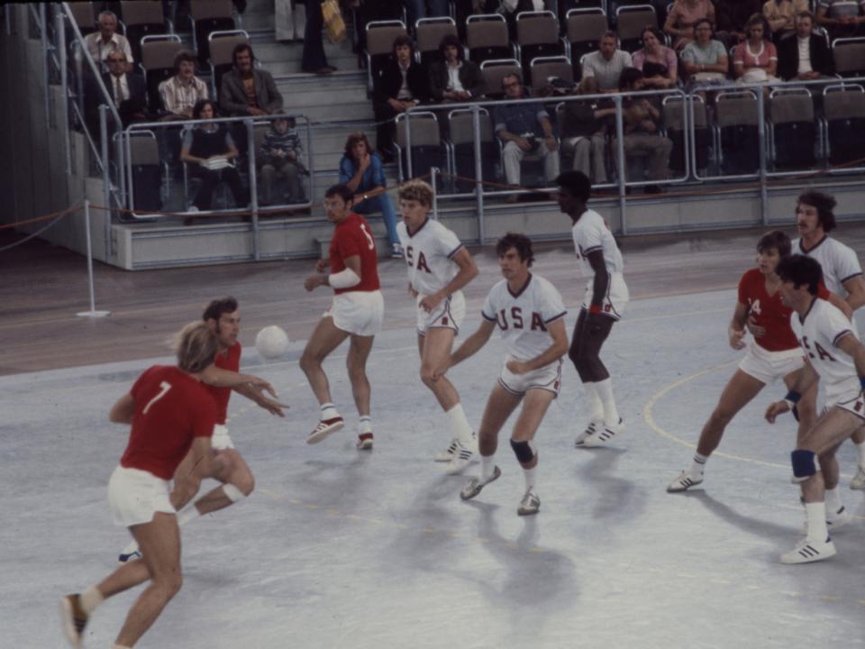 A wide shot of a group of men playing handball with an audience in the background.