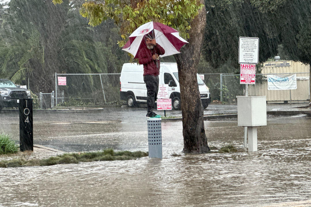 A man carrying an umbrella balances on a cylindrical metal post in the middle by a flooded street.