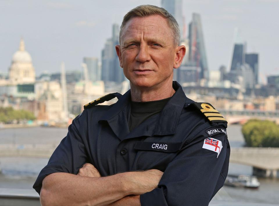 Daniel Craig wears a Royal Navy commander's uniform with his name on it as he crosses his arms with the London skyline behind him.