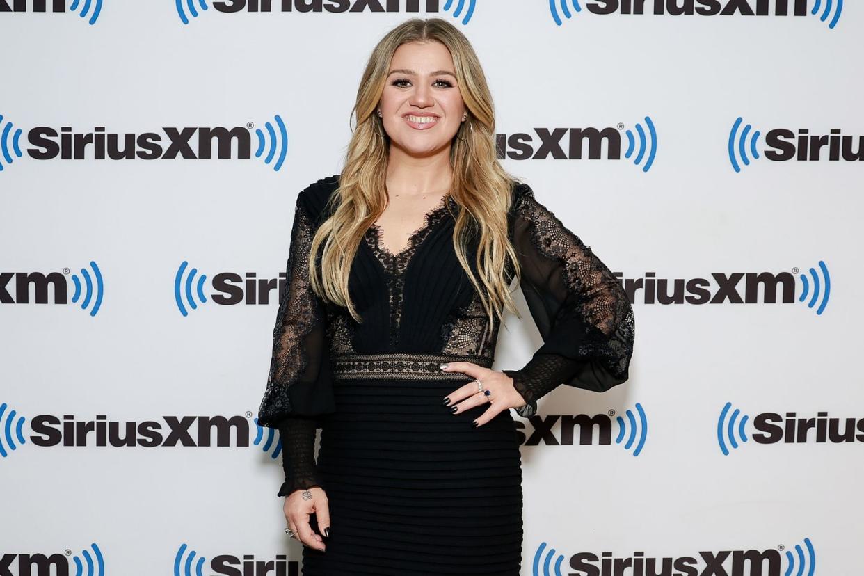 kelly clarkson posing with her hand on her waist, she's wearing a black dress with lacy sleeves and inserts on her midriff