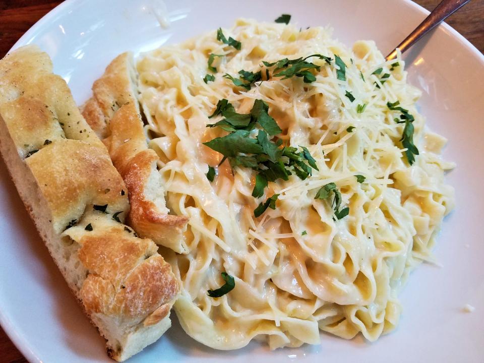Fettuccini Alfredo in a white bowl topped with parsley and cheese. There is also a piece of bread in the bowl and a spoon.