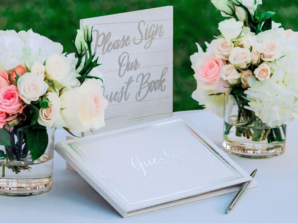 White guestbook with sign saying "Please sign our guest book" with pink and white flowers on either side