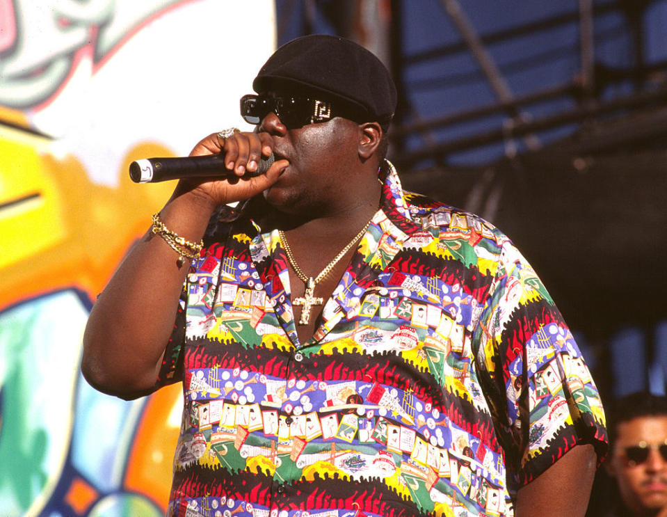 The image shows The Notorious B.I.G. performing on stage, holding a microphone and wearing a colorful graphic shirt, sunglasses, a black hat, and gold jewelry