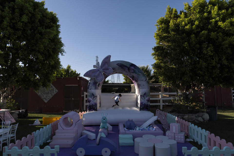 Max Cortes, 2, plays in an inflatable bouncer while waiting for a baptism ceremony to start at Rancho El Refugio date palm ranch owned by Carlos Ulloa in Twentynine Palms, Calif., Saturday, June 15, 2024. Ulloa's vision was to create places where he could keep his horses and have a working ranch with sheep and peacocks, while hosting events where a girl celebrating her quinceañera could ride in a horse-drawn carriage and families could have ample space to invite their relatives to a celebration without going broke. (AP Photo/Jae C. Hong)